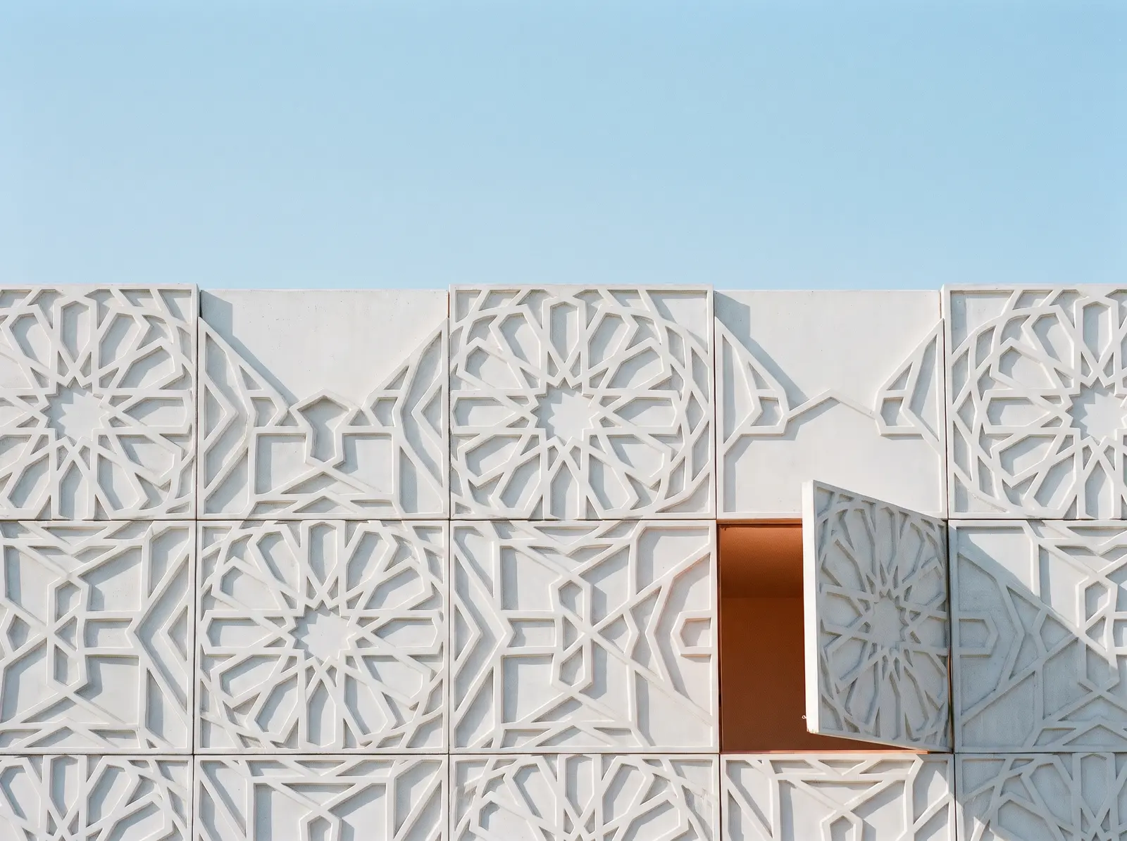 A white facade of carved geometric star tiles against a clear blue sky, one panel swung open onto an orange wall
